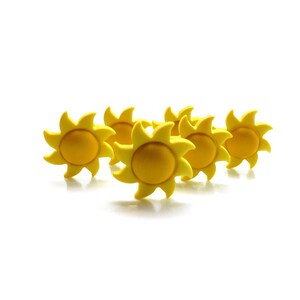 Puede incluir: Seis botones decorativos amarillos con forma de sol. Cada botón presenta un círculo central en relieve rodeado de rayos de sol estilizados. Los botones están dispuestos sobre un fondo blanco, creando una estética brillante y alegre.