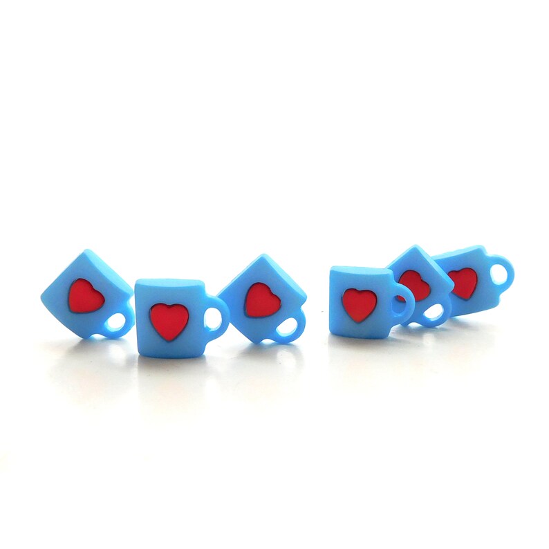 May include: A collection of light blue miniature coffee cup charms, each featuring a red heart design. The charms are arranged in a row, with some tilted at an angle. The background is plain white.