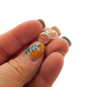 Puede incluir: Tres cuentas de vidrio transparente e iridiscente de diferentes tamaños, sostenidas en una mano con uñas pintadas.