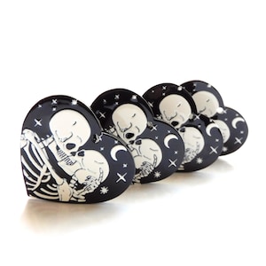 Puede incluir: Cinco pines de esmalte en forma de corazón negro con un diseño de pareja de esqueletos blancos besándose. El fondo presenta un cielo nocturno estrellado negro con una luna creciente.