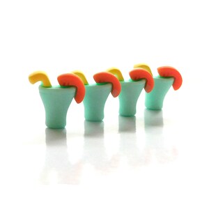 May include: Four miniature turquoise margarita glasses, each with a yellow straw and an orange citrus wedge. The drinks are arranged in a row against a white background, creating a playful and colorful display.