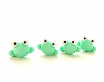 Puede incluir: Cuatro pequeñas figuras con forma de rana en color verde menta, hechas de un material liso. Cada rana presenta dos grandes ojos blancos saltones con pupilas negras y una línea curva grabada que representa una boca sonriente.