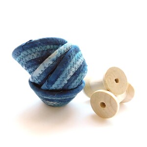 May include: Three small, round, blue fabric bowls with a stitched design are stacked together. Next to the bowls are three wooden spools. The bowls have a gradient of blue hues, and the spools are a natural wood color.