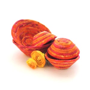 May include: A collection of handmade, colorful fabric bowls in shades of orange, pink, and yellow. The bowls are nested together, showcasing a spiral design. Two yellow roses are placed near the bowls, adding a touch of nature.