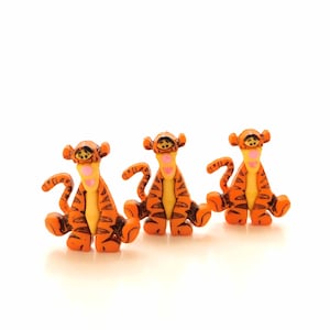 Peut inclure: Trois figurines de tigre orange et noir avec des rayures noires et des nez roses. Les figurines sont assises droites, la queue enroulée derrière elles.