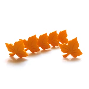 May include: Six orange maple leaf-shaped decorative items. The leaves are arranged in a row, with one leaf slightly separated from the others. The leaves have detailed veining and a smooth, matte finish. The background is plain white.