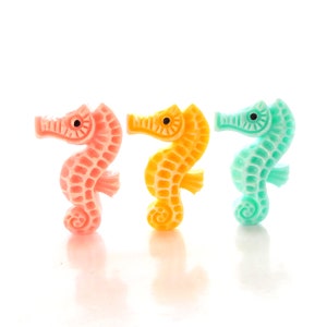 May include: Three small plastic seahorse figurines in pink, yellow, and blue. The seahorses are facing the right side of the image.
