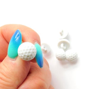 May include: A collection of small, white golf ball-shaped buttons. The buttons have a textured surface with dimples, resembling miniature golf balls. Several buttons are scattered on a white surface.