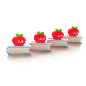 May include: Four miniature red apple bookends with a green leaf and the text "A+" on each apple. The bookends are on a white surface.