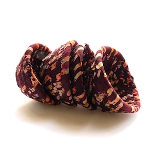 May include: A fabric scrunchie in a spiral shape, featuring a pattern of burgundy, orange, and cream-colored shapes. The scrunchie is made of a textured material and is set against a white background.