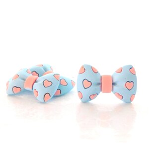 May include: Four light blue bow-shaped hair clips with pink hearts printed on them. The bows are arranged in a pile.