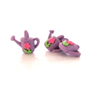 May include: A collection of small, purple watering can-shaped embellishments. Each can features a pink flower with green leaves. The watering cans are made of a smooth material and are designed with a handle and a spout.