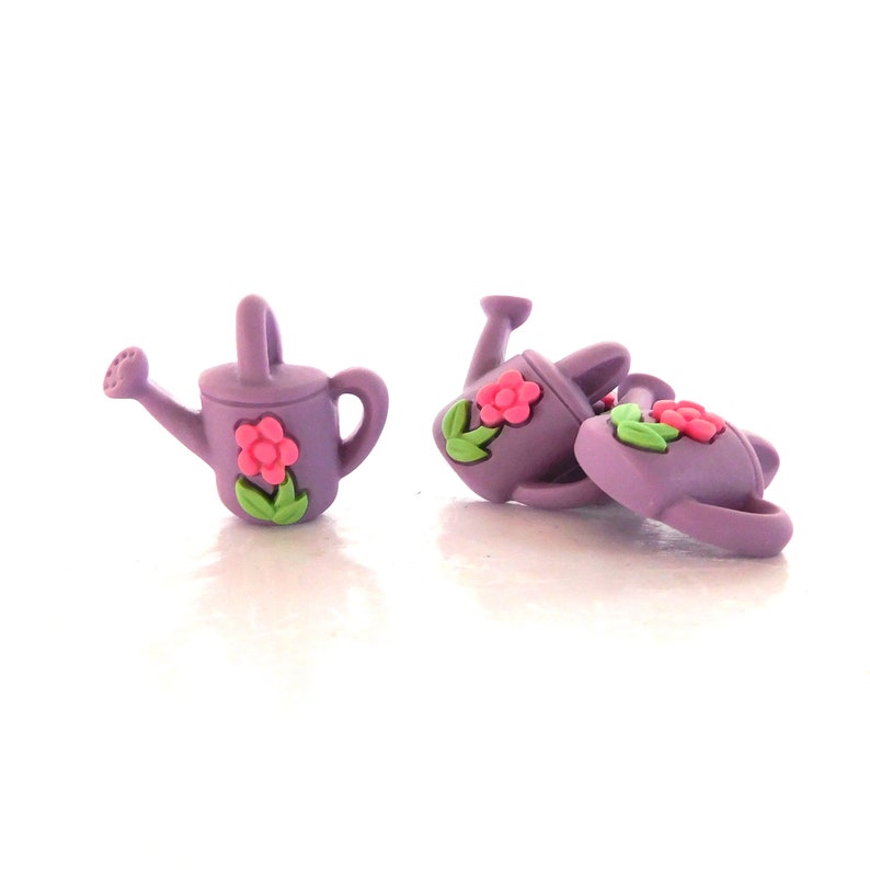 May include: A collection of small, purple watering can-shaped embellishments. Each can features a pink flower with green leaves. The watering cans are made of a smooth material and are designed with a handle and a spout.