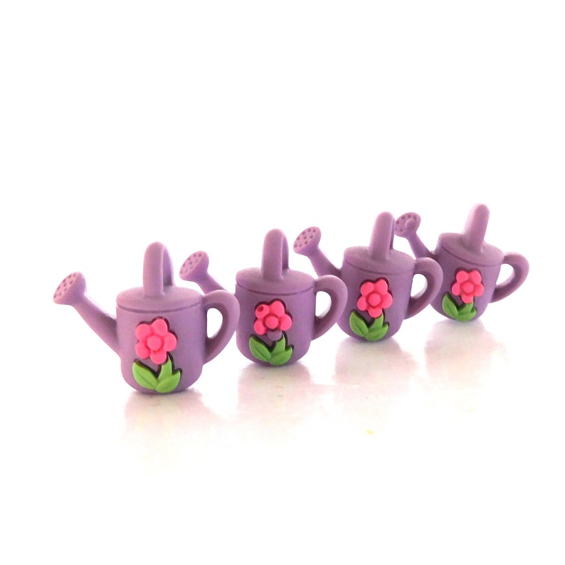May include: Four lavender watering can-shaped buttons with pink flower and green leaf accents. Each button has a handle and a spout. The buttons are arranged in a row against a white background.