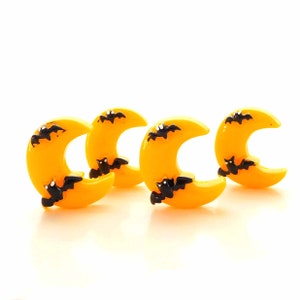May include: Four yellow crescent moon-shaped decorations with black bat silhouettes. These Halloween-themed ornaments are arranged on a white surface, creating a festive and spooky display.