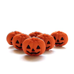 May include: Six small, orange pumpkin decorations with black carved faces. The pumpkins are arranged on a white surface, perfect for Halloween or fall-themed crafts and decor.