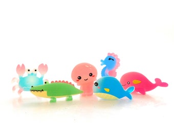 May include: A collection of six small, colourful animal figurines including a light blue crab, a green crocodile, a pink octopus, a blue seahorse, a blue whale, and a pink dolphin, all featuring smooth surfaces and simple, friendly facial expressions.