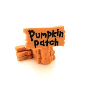 以下が含まれることがあります： オレンジ色の背景に黒い文字で「パンプキンパッチ」と書かれた小さな木製の看板。看板は木製のブロックの積み重ねの上に置かれている。