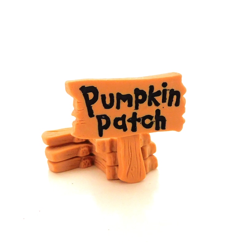 K&ouml;nnte beinhalten: Ein kleines Holzschild mit den Worten "Pumpkin Patch" in Schwarz auf orangefarbenem Hintergrund. Das Schild steht auf einem Stapel Holzkl&ouml;tzen.