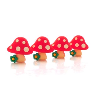 May include: Four mushroom-shaped ornaments. Each mushroom features a red cap with white spots, a beige stem, and a green flower with a yellow centre at the base. They are arranged in a row against a white background.