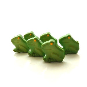 May include: Five green frog figurines with orange eyes. The frogs are arranged in a line, facing the same direction.