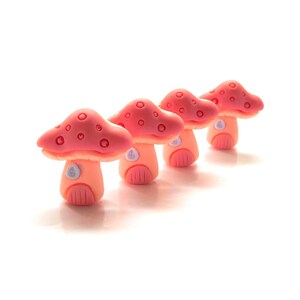 May include: Four miniature mushroom figurines in a row. Each mushroom has a pink cap with small, round indentations and a peach-colored stem with a door and window design. The background is white.