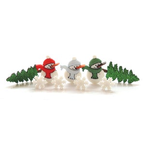 May include: Three small snowman figurines with orange carrot noses and black dotted smiles. Each wears a hat and scarf in red, light blue, or green. They are arranged with four white plastic snowflakes and two glittery green pine trees.