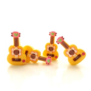 May include: Four yellow guitar-shaped buttons featuring a pink headstock with a green heart detail, a brown fretboard, and a white flower design around the soundhole. Each button has a small pink accent near the base of the guitar body.