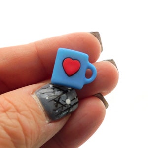May include: A small, light blue mug charm with a red heart in the center. The mug has a handle and is made of a smooth material. The charm is a cute, miniature representation of a coffee cup.