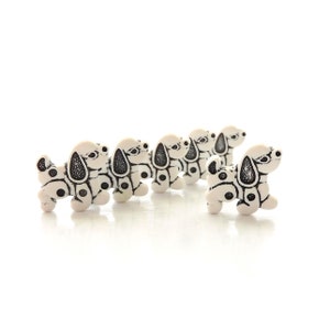 May include: A row of six small, white dog figurines with black spots and black ear markings. The figurines are arranged in a line, with one slightly offset from the others. The dogs are standing on a white surface.