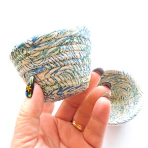 May include: Two small, round bowls constructed from coiled fabric strips featuring a marbled pattern in shades of blue, green, and white. The fabric coils are joined together with visible white zigzag stitching throughout the exterior and interior.