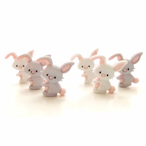 May include: A collection of six small rabbit figurines arranged in a group. Each bunny features a smooth finish, upright ears with pink inner details, a small pink nose, and a round pink tail, rendered in alternating shades of white and gray.