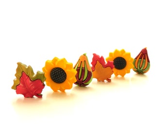 May include: A collection of six decorative buttons shaped like autumn elements, including red and green maple leaves, yellow sunflowers with dark centers, and multi-colored gourds in shades of orange, green, and deep red, arranged in a row.