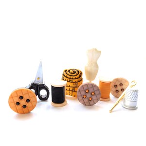 Peut inclure: Une collection de breloques miniatures sur le thème de la couture comprenant un bouton jaune, des ciseaux noirs, une bobine de fil noire, un mètre ruban jaune, un mannequin, un bouton marron, une bobine orange, un bouton beige et un dé à coudre.