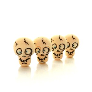 May include: Four small, off-white skull-shaped objects with black outlines and blue eyes are arranged in a row. Each skull has a jagged line on top and a stitched mouth. The objects are likely decorative or for crafting.