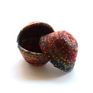 May include: A small, round, lidded basket with a colorful, textured exterior. The basket is open, revealing a dark interior. The exterior features a pattern of red, blue, and yellow dots and stripes.