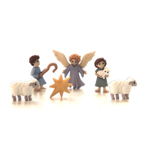 May include: A nativity scene set with a blue-winged angel, two shepherds, a star, and two white lambs. The angel is wearing a lavender robe and has blonde hair. The shepherds are wearing blue robes and have brown hair. The star is yellow and has five points. The lambs are white and have black eyes.