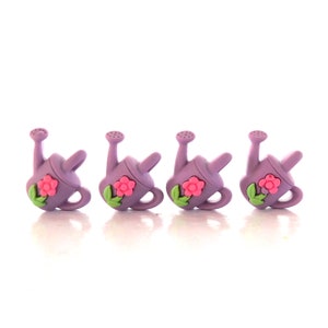 May include: Four lavender watering can charms with pink flowers and green leaves. The watering cans have a handle and a spout. The charms are arranged in a row against a white background.
