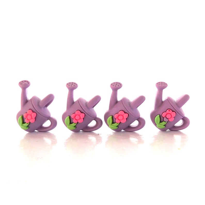 May include: Four lavender watering can charms with pink flowers and green leaves. The watering cans have a handle and a spout. The charms are arranged in a row against a white background.