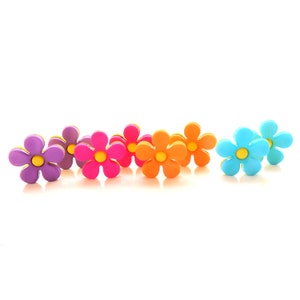 May include: A collection of colorful flower-shaped decorative buttons. The buttons are in various colors, including purple, pink, orange, and blue, each with a yellow center. The buttons are arranged on a white surface.