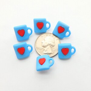 May include: Six small, light blue mug-shaped charms, each with a red heart, are arranged around a quarter. The charms have a handle on the side. The background is white.