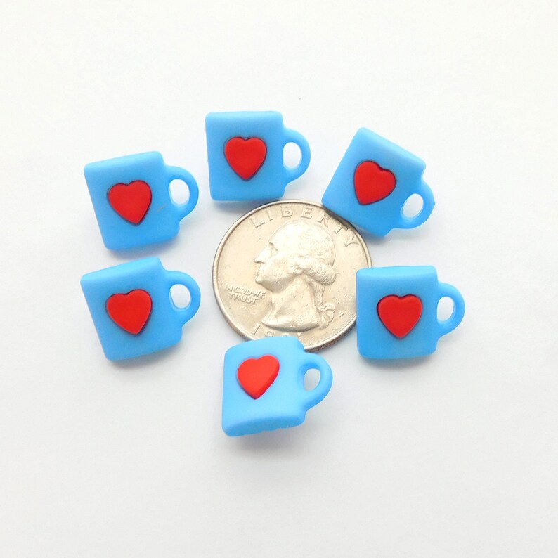 May include: Six small, light blue mug-shaped charms, each with a red heart, are arranged around a quarter. The charms have a handle on the side. The background is white.
