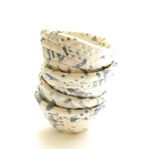 May include: A stack of five small, handmade fabric bowls. The bowls are cream-colored with a blue and gray speckled pattern. The bowls are stacked on top of each other, with the top bowl slightly angled.