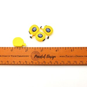 K&ouml;nnte beinhalten: Vier gelbe Messband-Anh&auml;nger mit dem Text "5m" auf jedem. Die Anh&auml;nger befinden sich auf einem Holzlineal mit dem Text "Button Up Your Creativity" und "Pieceful Design" darauf.
