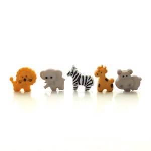 May include: A set of five small animal-shaped buttons arranged in a row. The collection includes an orange lion, a light gray elephant, a white zebra with black stripes, an orange giraffe with brown spots, and a light gray hippopotamus.