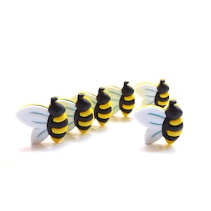 May include: A collection of five small bee-shaped buttons. Each button features a black head, a yellow and black striped body, and two white wings with light blue accent lines. The buttons are arranged in a cluster against a plain white background.