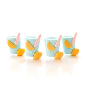 May include: Four miniature blue ceramic cups with yellow lemon slices and pink straws. The cups are arranged in a row on a white surface.