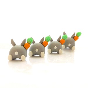 Puede incluir: Cuatro figuritas de conejo grises con pies blancos y una zanahoria en la espalda. Las zanahorias son naranjas con puntas verdes.