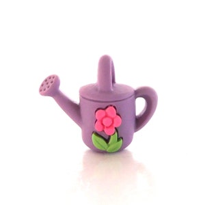 May include: A small, lavender watering can with a pink flower and green leaves. The watering can has a handle and a spout with small holes. The flower is in the center of the can.