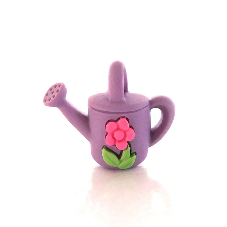 May include: A small, lavender watering can with a pink flower and green leaves. The watering can has a handle and a spout with small holes. The flower is in the center of the can.